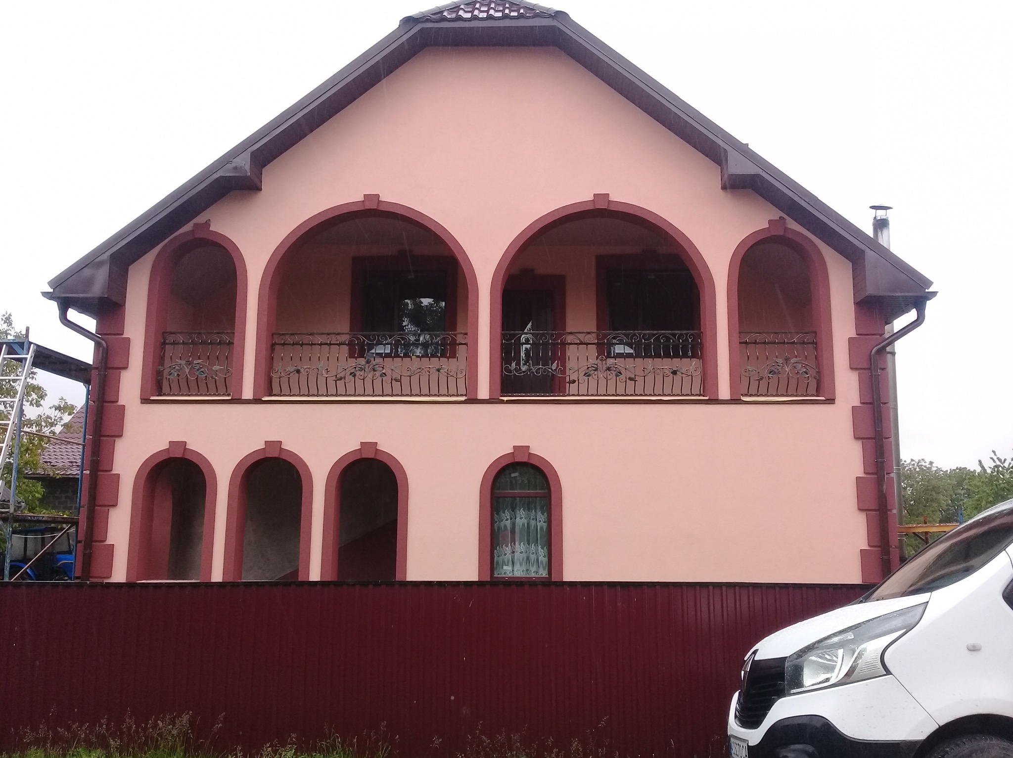 Dwukondygnacyjny dom z balkonem i łukowymi oknami, pomalowany na różowo z bordowymi akcentami. Widoczny fragment białego samochodu i drabina z boku budynku.