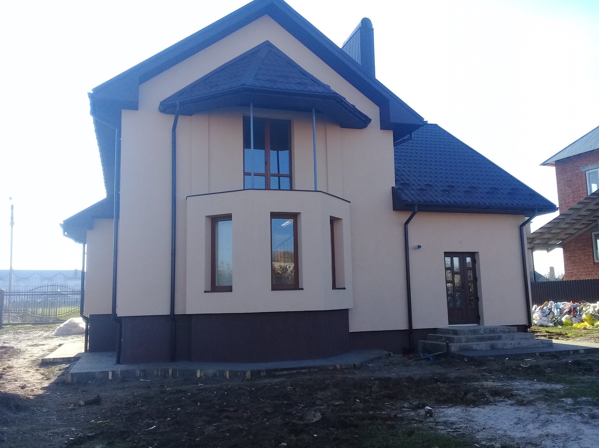 Elewacja domu jednorodzinnego w beżowej tonacji z ciemną podmurówką i dachem. Widoczne okna z drewnianymi ramami i wejście z betonowymi schodami. Teren wokół domu częściowo zagospodarowany.