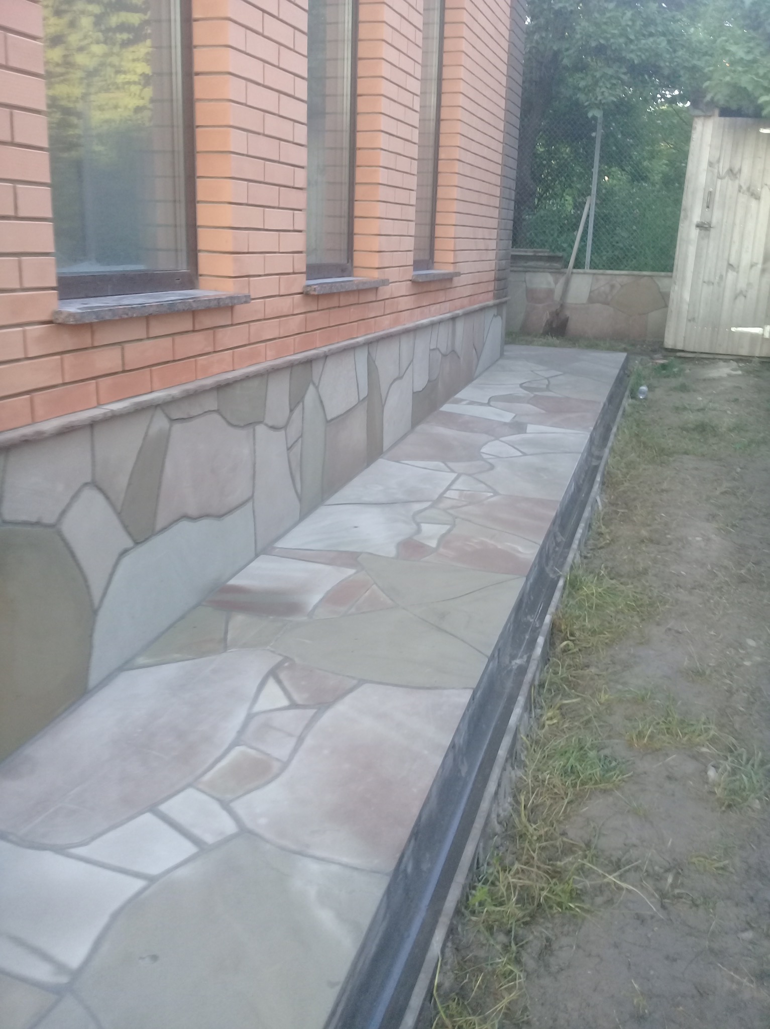 Wykończony fragment elewacji budynku z dekoracyjnym kamieniem i płytami chodnikowymi w odcieniach beżu i szarości, widok z zewnątrz, ceglana ściana z oknami w tle.