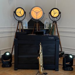 Damian Brzeziński - Scena z saksofonem na statywie, czarnym DJ-skim stanowiskiem Humper i trzema reflektorami na drewnianych trójnogach, oświetlającymi wnętrze w ciepłych barwach.