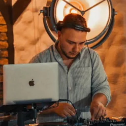 DJ z laptopem Pioneer DJ w Otwocku, grający muzykę na imprezie. Duża lampa oświetleniowa w tle. Mężczyzna w słuchawkach, skupiony na miksowaniu.