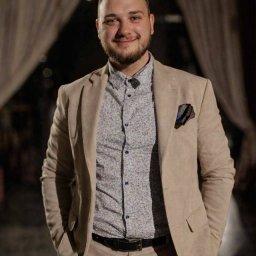 Damian Brzeziński - Uśmiechnięty mężczyzna w beżowym garniturze i wzorzystej koszuli, z chusteczką w kieszonce, pozuje na tle ciemnego wnętrza z dekoracyjnymi zasłonami.