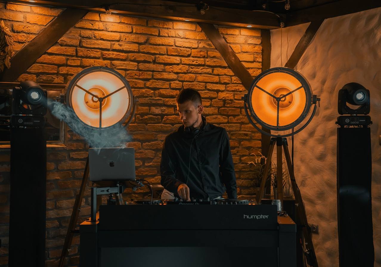 DJ w czarnej koszuli i słuchawkach miksuje muzykę na konsoli Pioneer DJ, otoczony ciepłym światłem reflektorów i dymem, z laptopem Apple w tle, na tle ceglanej ściany i drewnianych belek.