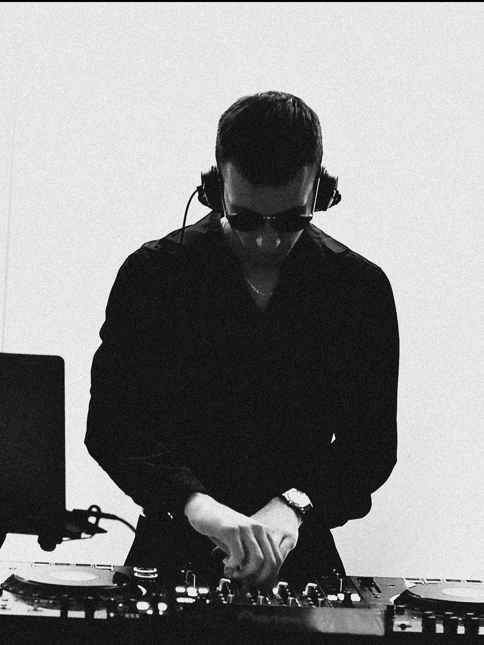 Czarno-białe zdjęcie DJ-a w słuchawkach i okularach przeciwsłonecznych, miksującego muzykę na profesjonalnym sprzęcie. Widoczny laptop i zegarek na ręce.