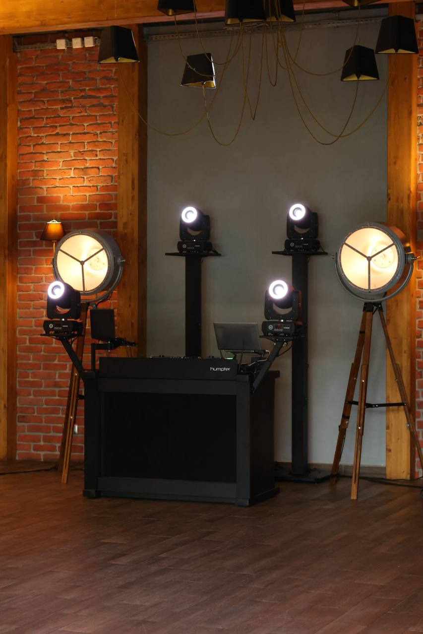 Nowoczesne stanowisko DJ'skie z oświetleniem scenicznym w loftowym wnętrzu z ceglaną ścianą i drewnianymi elementami konstrukcyjnymi.