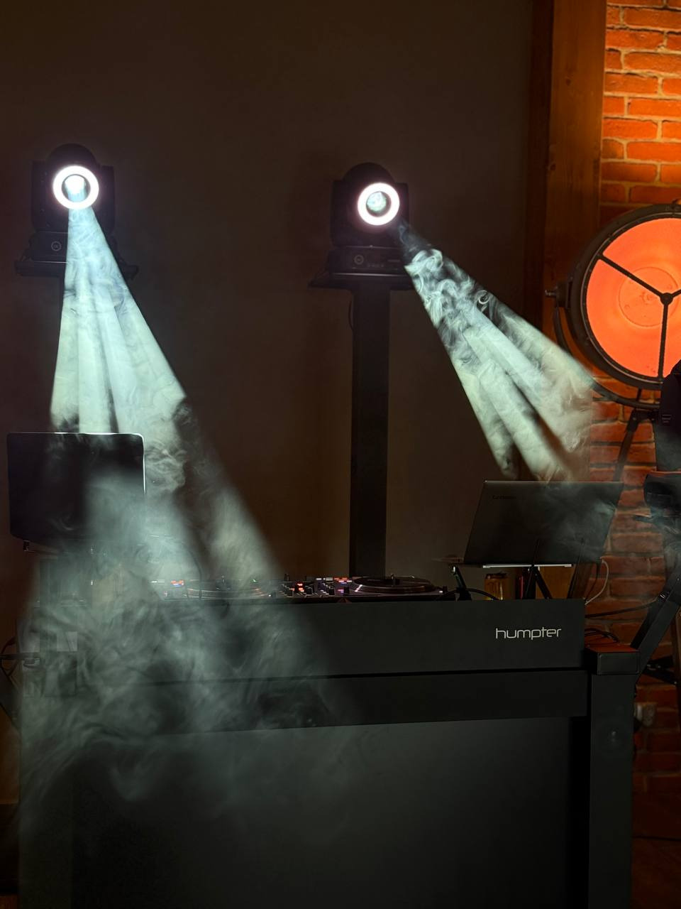 Stanowisko DJ-a z oświetleniem scenicznym i dymem. Dwa reflektory oświetlają konsoletę, a w tle widoczna ceglana ściana i lampa.