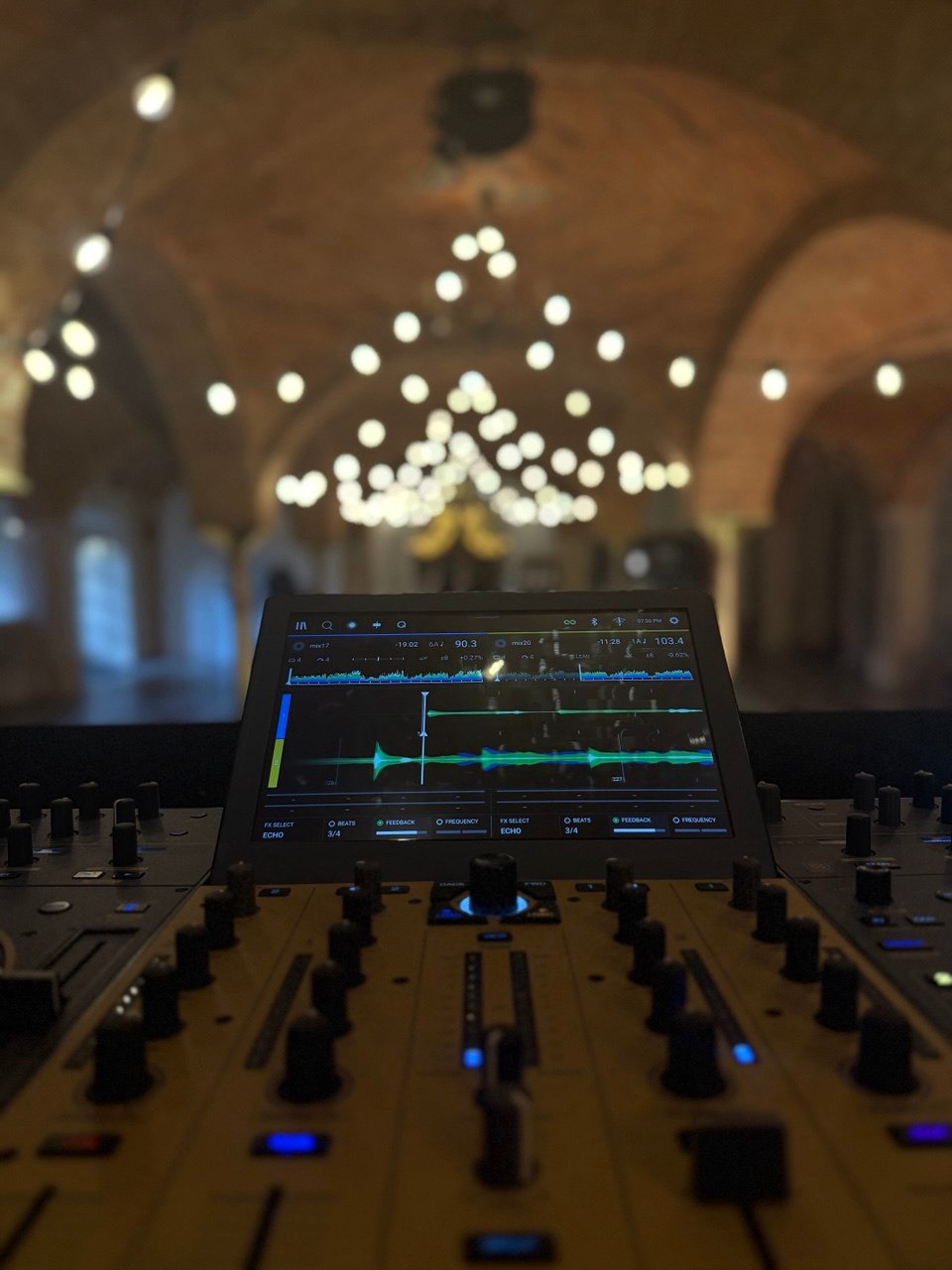 Konsoleta DJ-ska z ekranem i suwakami na tle sali z oświetleniem punktowym, ustawiona pod kątem, ukazująca instrumenty pracy DJ-a.