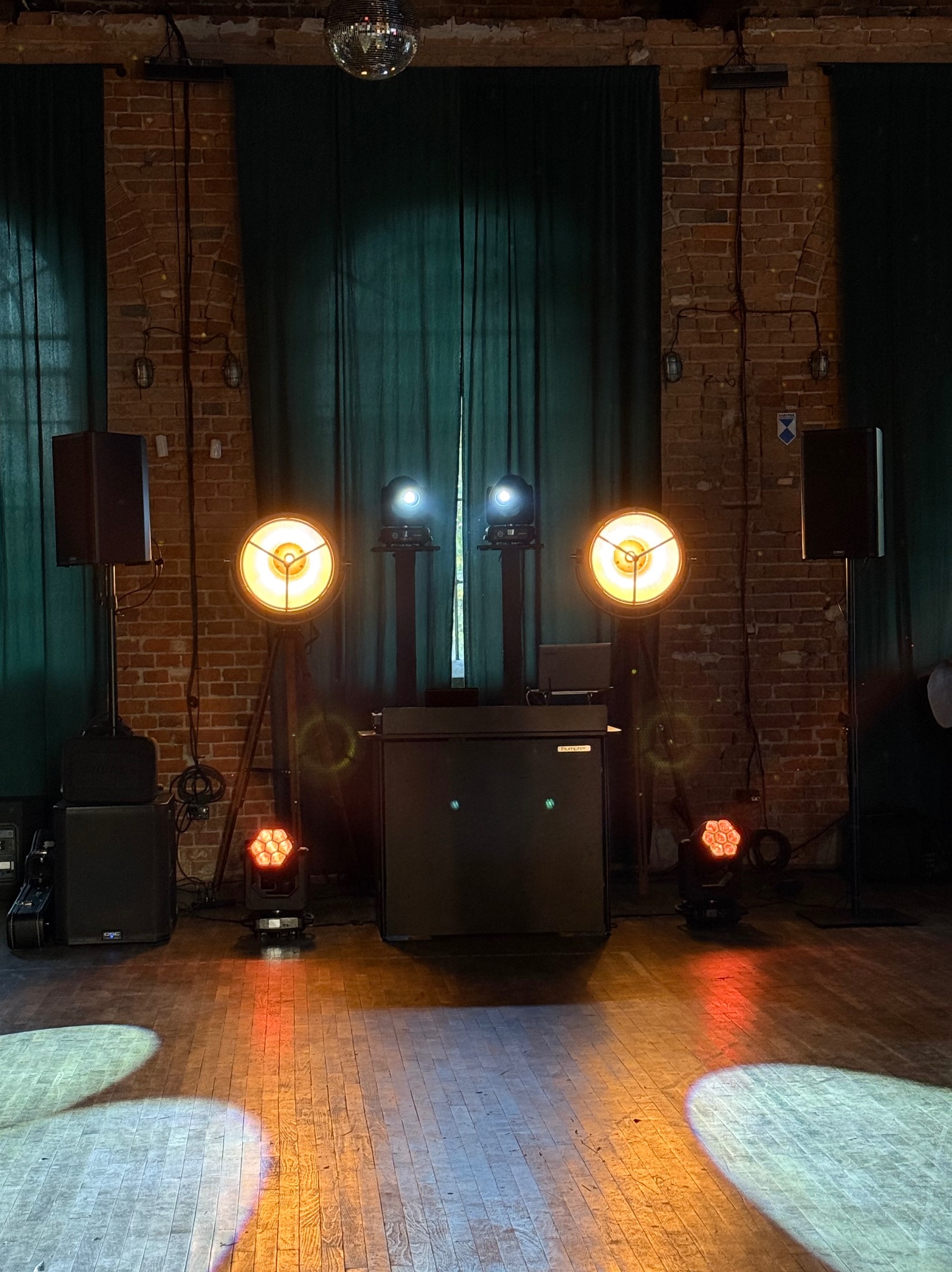 Sprzęt nagłośnieniowy i oświetleniowy w sali z ceglanymi ścianami i drewnianą podłogą. Dwa reflektory na statywach, kolumny głośnikowe, kula dyskotekowa.