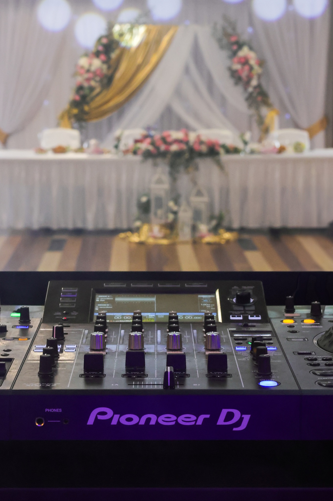 Konsoleta DJ marki Pioneer na tle dekoracji weselnej z kwiatami i białymi zasłonami, oświetlona punktowo. Widoczne pokrętła i przyciski.
