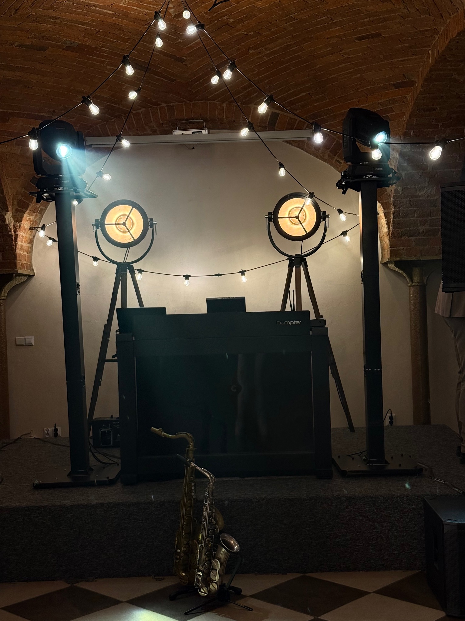 Scena z oświetleniem eventowym i saksofonem na parkiecie. Dwa reflektory, dekoracyjne żarówki i sprzęt DJ-ski tworzą klimat gotowy na występ muzyczny.