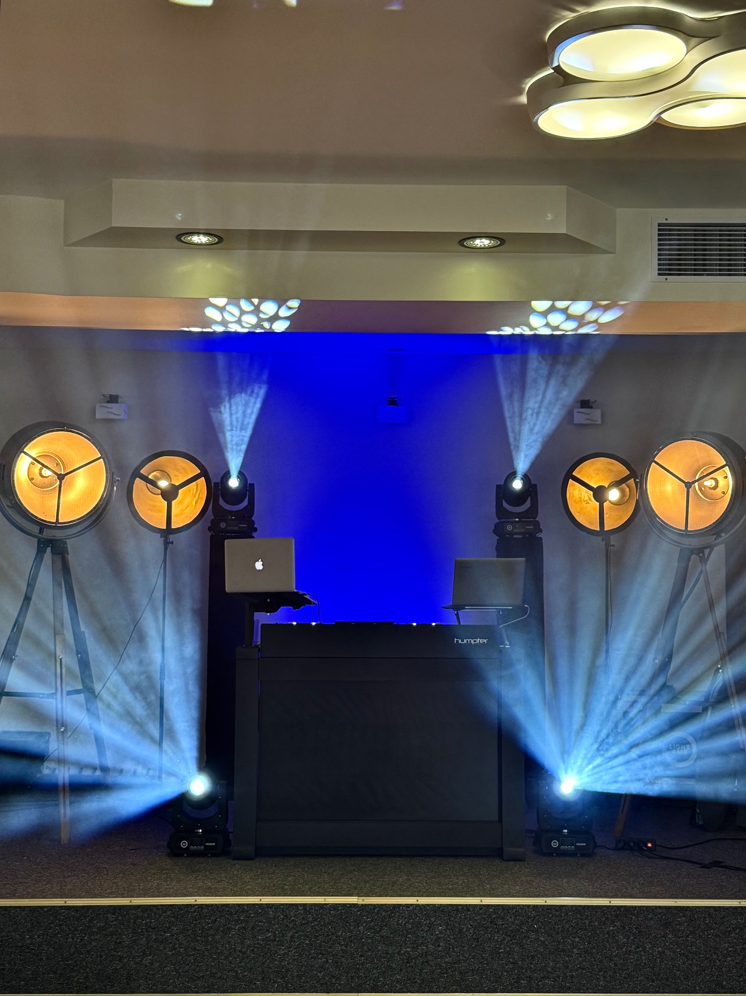 Scena z profesjonalnym oświetleniem eventowym: reflektory na trójnogach, ruchome głowy, konsoleta DJ-ska z laptopami, tworzące dynamiczną atmosferę światłem.
