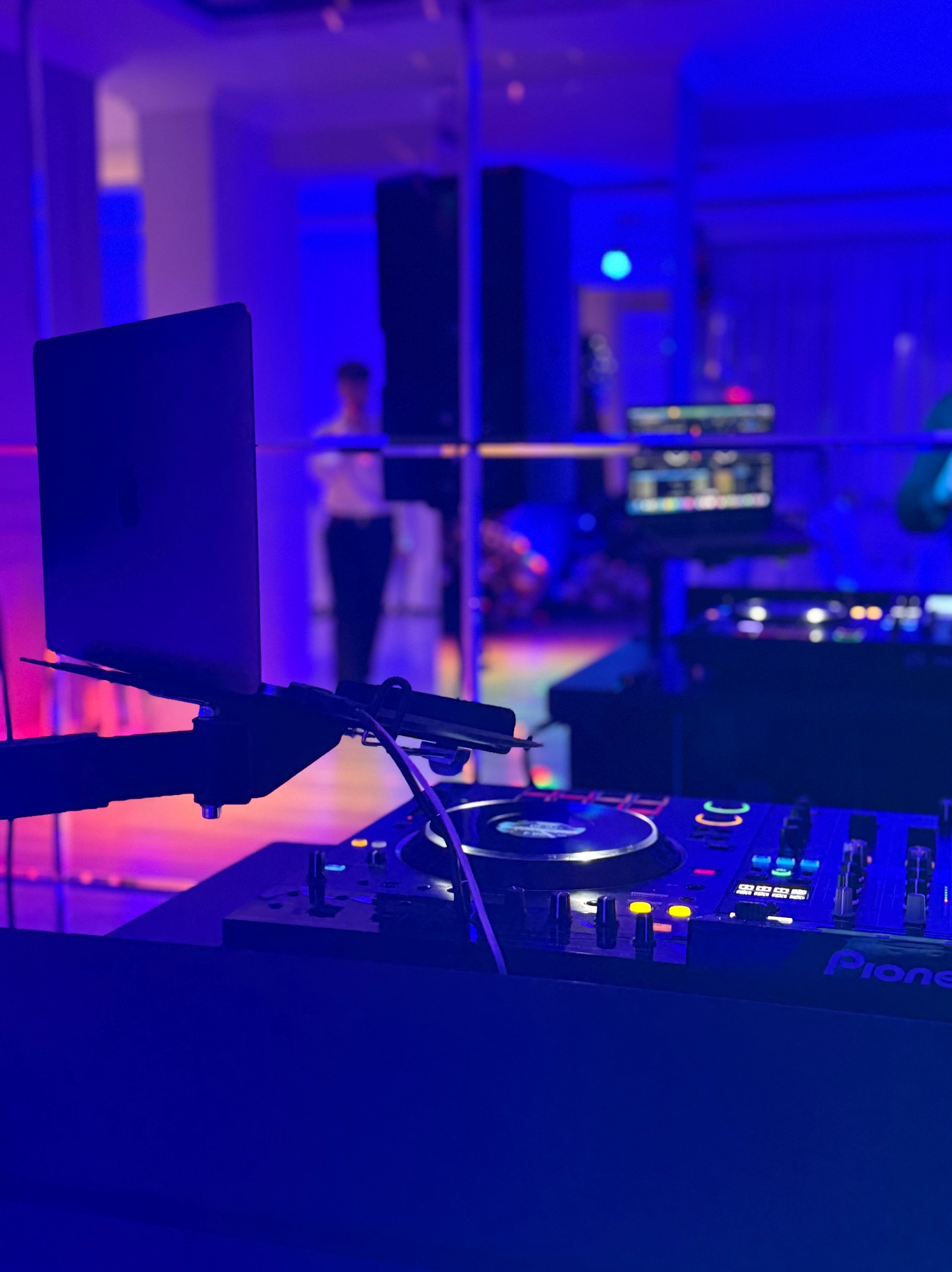 Konsoleta DJ Pioneer z laptopem w tle sali eventowej oświetlonej na niebiesko, widoczna postać w oddali. Sprzęt nagłośnieniowy na imprezę.