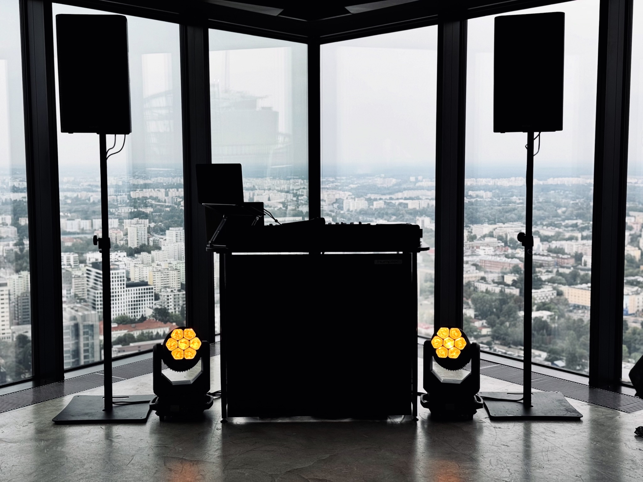 DJ stanowisko z nagłośnieniem i oświetleniem scenicznym na tle panoramy miasta za oknem. Sprzęt gotowy do występu, ustawiony w rogu pomieszczenia.