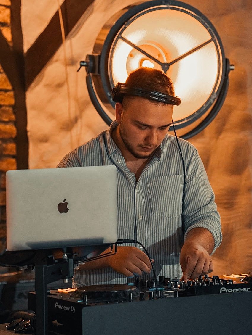 DJ z laptopem Pioneer DJ w Otwocku, grający muzykę na imprezie. Duża lampa oświetleniowa w tle. Mężczyzna w słuchawkach, skupiony na miksowaniu.