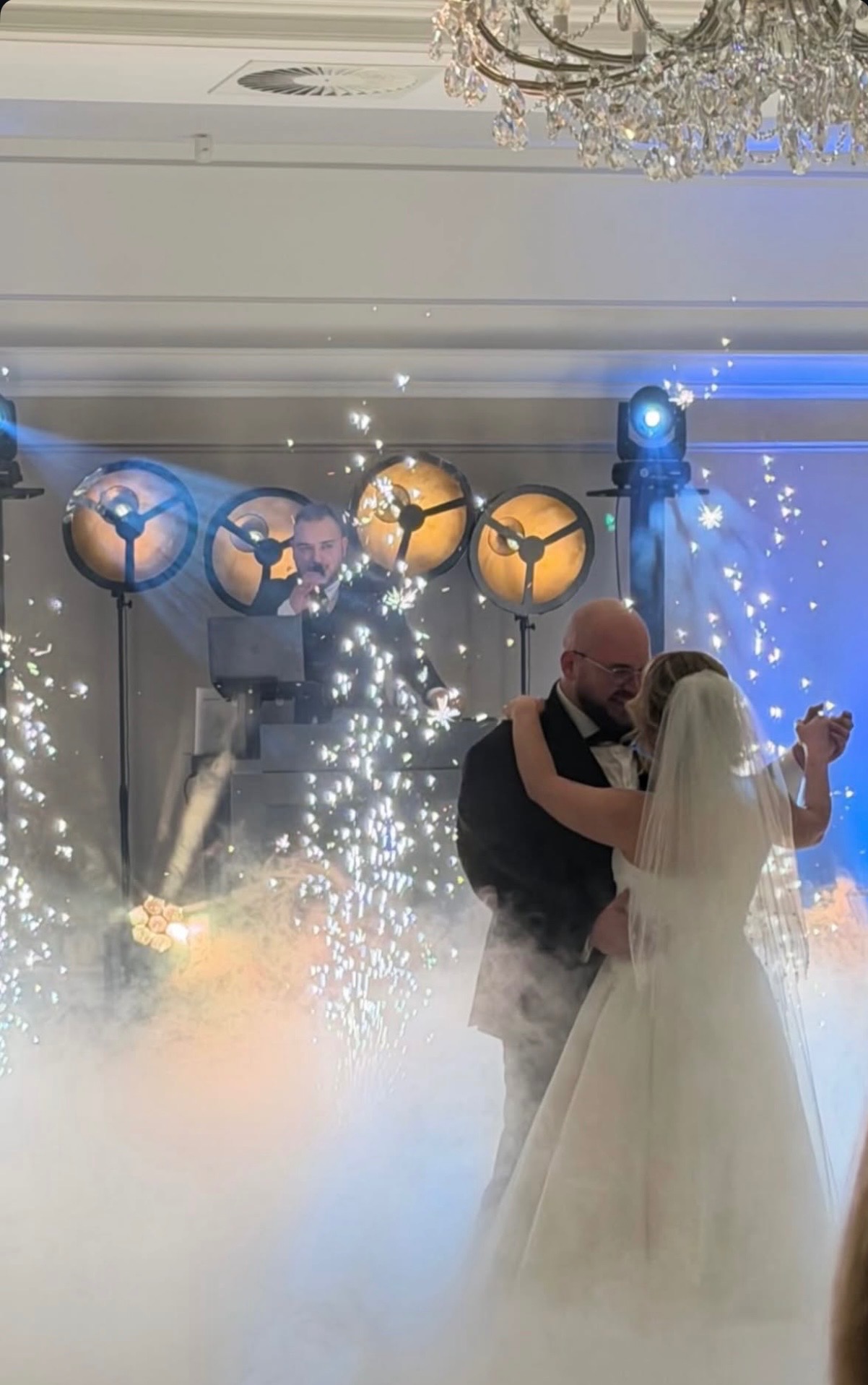 Para młoda tańczy w dymie i blasku iskier, za nimi DJ z oświetleniem na weselu. Dekoracja sali z żyrandolem.