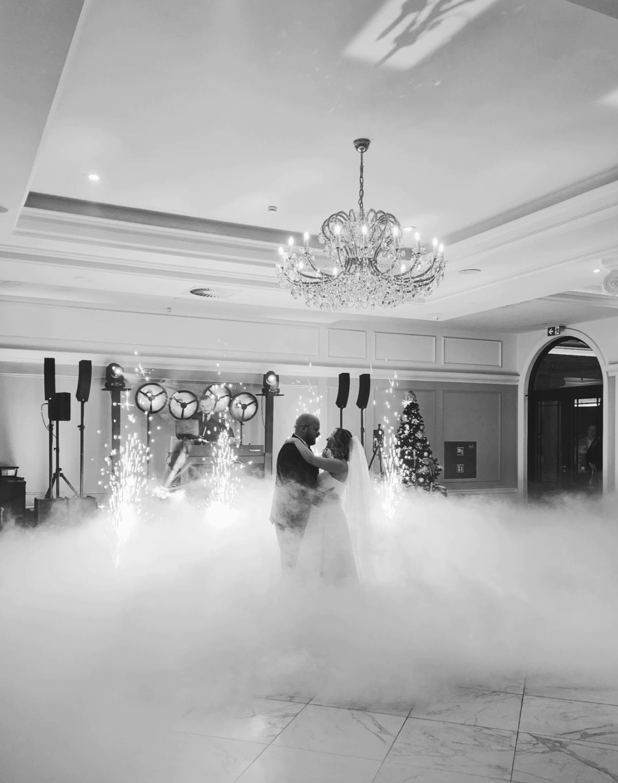 Czarno-białe zdjęcie pary młodej tańczącej w dymie, z fontannami iskier w tle i oświetleniem scenicznym. W tle choinka i żyrandol.