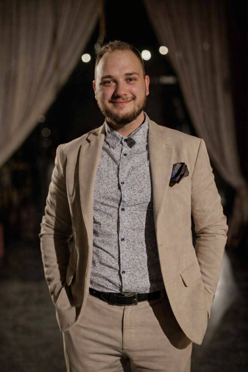 Uśmiechnięty mężczyzna w beżowym garniturze i wzorzystej koszuli, z chusteczką w kieszonce, pozuje na tle ciemnego wnętrza z dekoracyjnymi zasłonami.