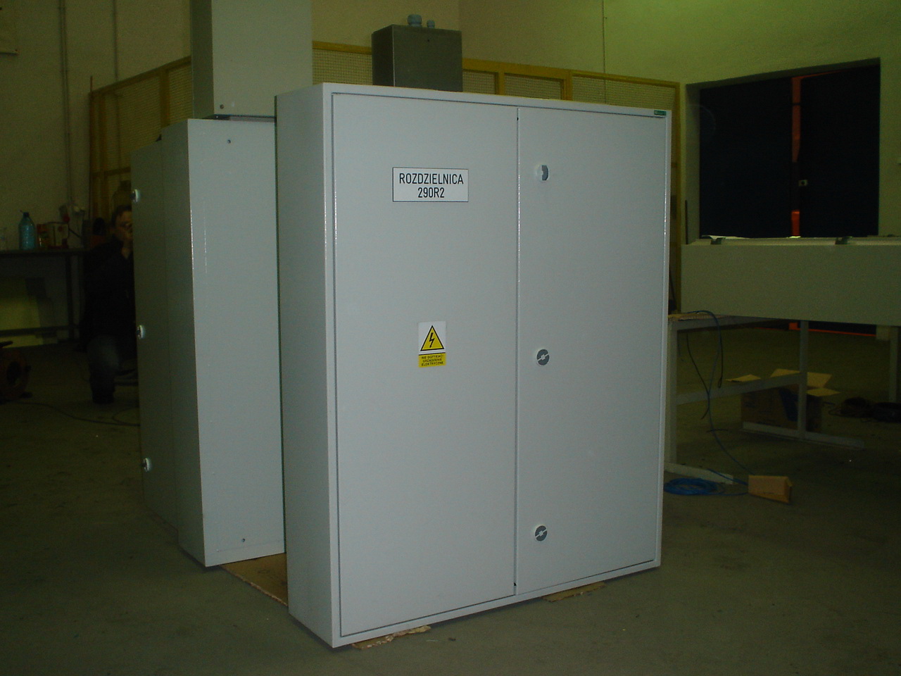 Dwie szare szafy elektryczne rozdzielcze z naklejką ostrzegawczą o zagrożeniu porażeniem elektrycznym, umieszczone w hali produkcyjnej, widoczne przewody i elementy wyposażenia.