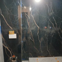 Grzegorz Diaków - Częściowo wyłożona czarnymi płytkami z marmurowym wzorem ściana łazienki, widoczne otwory na armaturę, gąbka na płytce i poziomowanie płytek na podłodze.