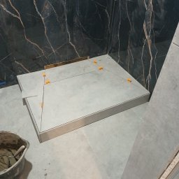 Grzegorz Diaków - Montaż płytek podłogowych w kabinie prysznicowej z użyciem poziomowania krzyżykami, widoczna ściana z ciemnego marmuru z jasnymi żyłami i wiadro z zaprawą.