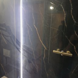 Grzegorz Diaków - Część wykończonej łazienki z czarnymi płytkami imitującymi marmur, podświetlona pionową listwą LED, widoczna bateria prysznicowa w kolorze złotym oraz fragment toalety.