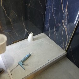 Grzegorz Diaków - Szczegółowe ujęcie montażu kabiny prysznicowej z użyciem silikonu, widoczna aplikator silikonu, biała ceramika i czarne marmurowe płytki na ścianie.