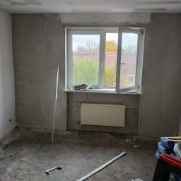 Grzegorz Diaków - Pokój w trakcie prac przygotowawczych do wykończenia: ściany pokryte szarą warstwą, widoczne narzędzia na parapecie i worki z materiałami budowlanymi na podłodze.