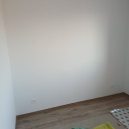 Grzegorz Diaków - Pomalowana na biało ściana w rogu pokoju z widocznym gniazdkiem elektrycznym, drewnianą listwą przypodłogową oraz fragmentem podłogi z panelami i zabezpieczeniem z folii malarskiej.