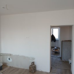 Grzegorz Diaków - Wnętrze w trakcie prac wykończeniowych, widoczna ściana z fragmentami nałożonej gładzi, instalacja elektryczna, podłoga zabezpieczona kartonem, oraz narzędzia i materiały budowlane w tle.