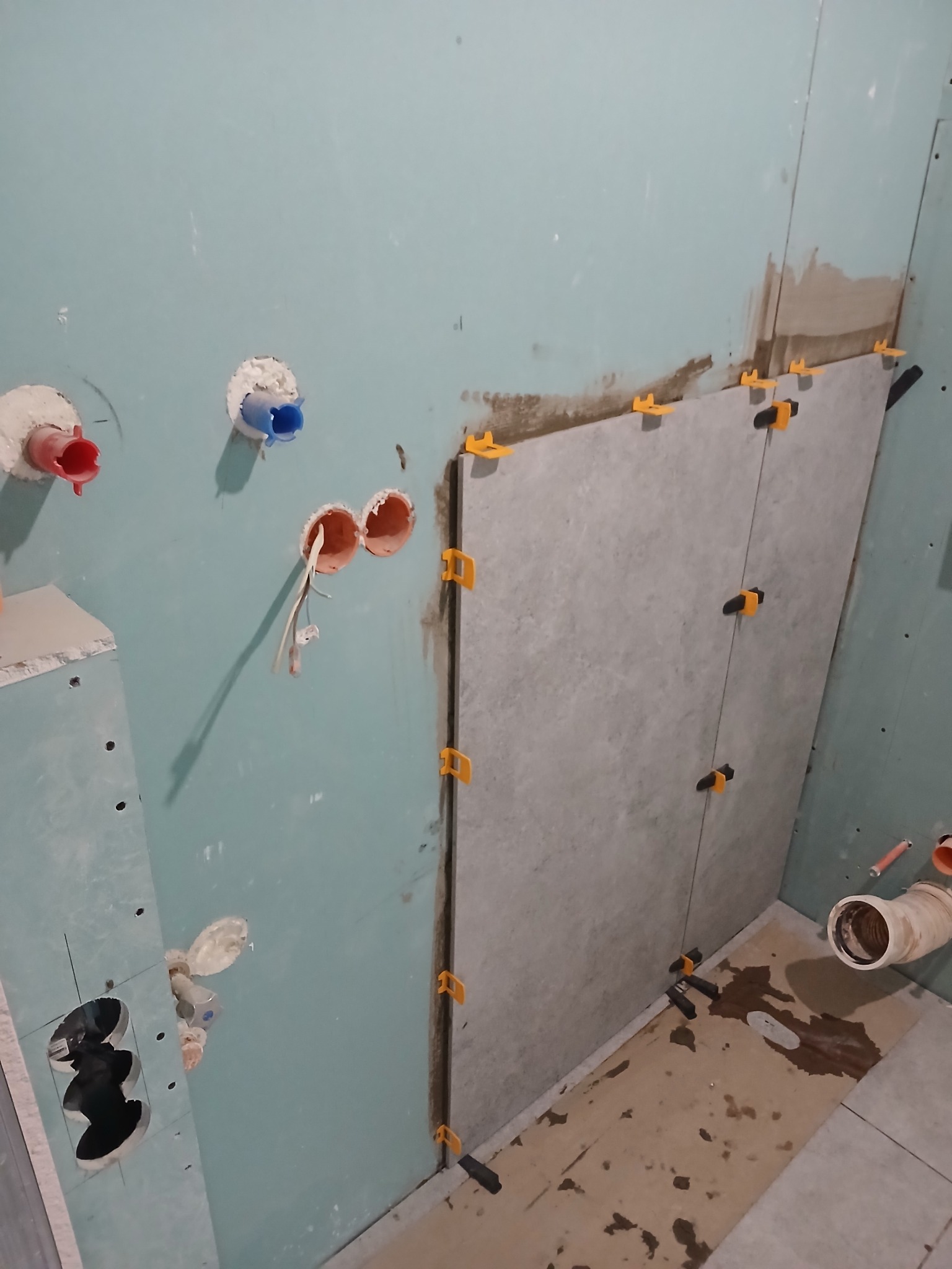 Świeżo przyklejona szara płytka na ścianie łazienki, z użyciem pomarańczowych klipsów poziomujących, widoczne wystające rury i kable, surowy stan wykończenia.