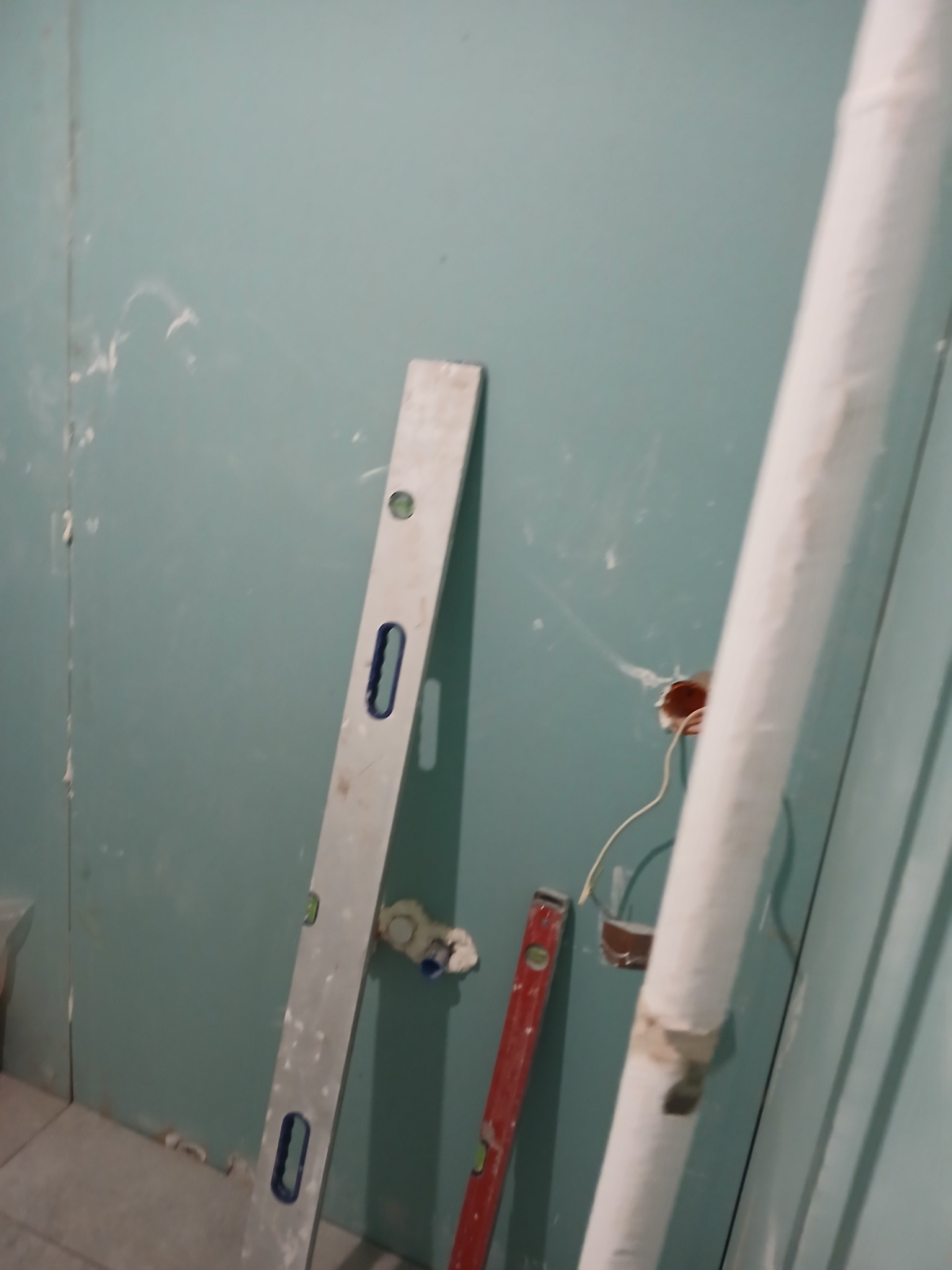 Ściana z płyty karton-gips w trakcie montażu, widoczne narzędzia: poziomica, puszka elektryczna z wystającymi przewodami, rura. Ślady po szpachlowaniu na ścianie.