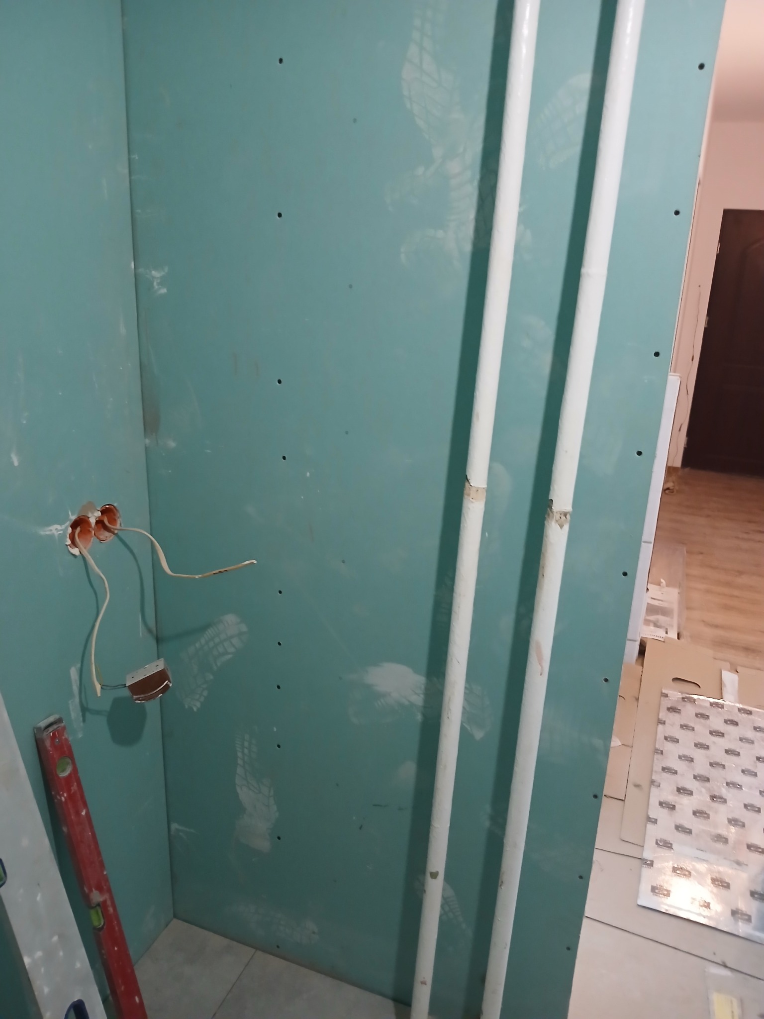 Ściana z zielonej płyty gipsowo-kartonowej, z widocznymi przewodami elektrycznymi wystającymi z puszki, pionowo ułożone rury oraz poziomica oparta o ścianę w rogu pomieszczenia.