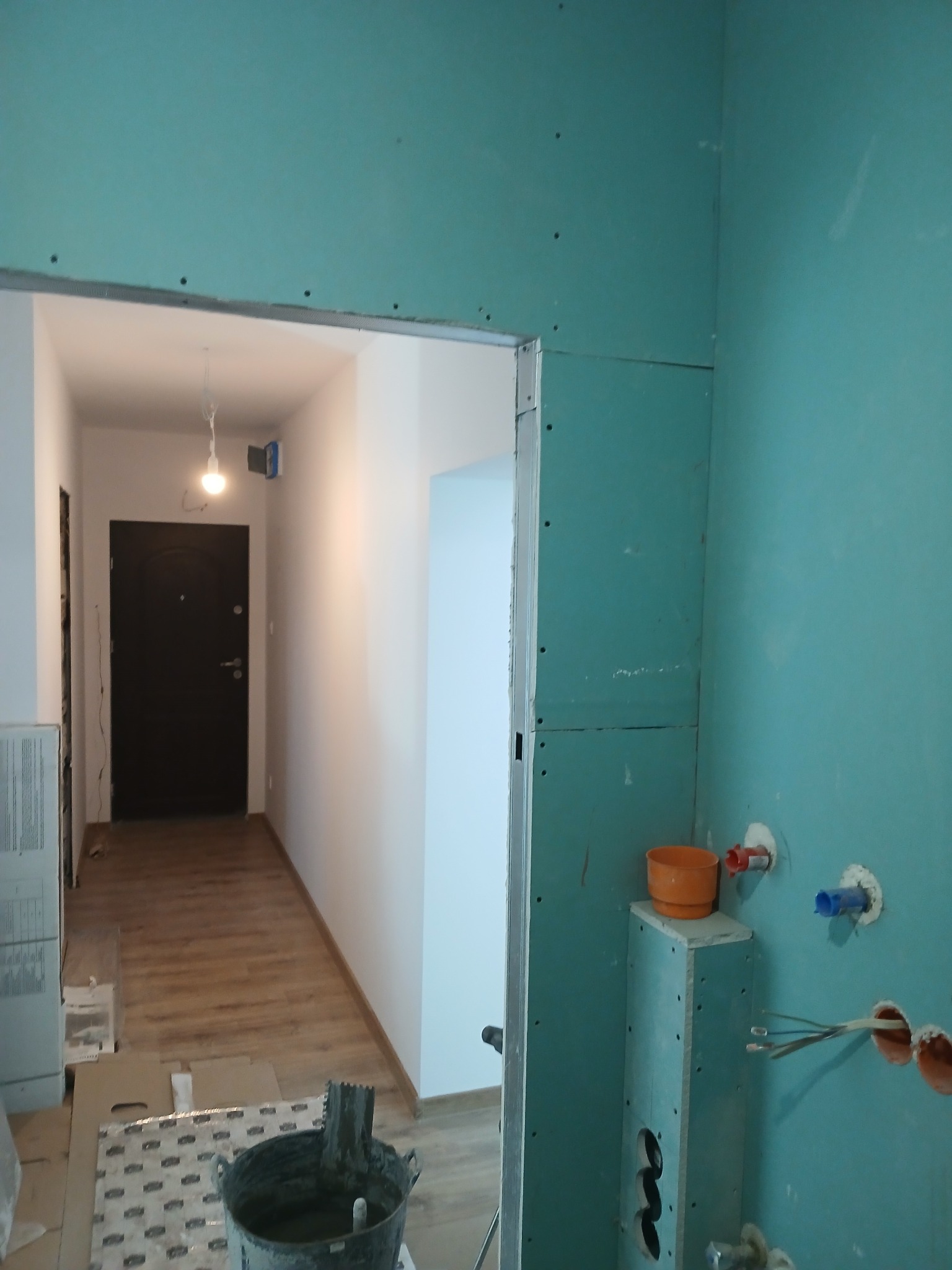 Wnętrze w trakcie prac wykończeniowych: ściany pokryte surową płytą gipsowo-kartonową w odcieniu turkusowym, widoczne instalacje wodne i elektryczne, fragment korytarza z ciemnymi drzwiami...