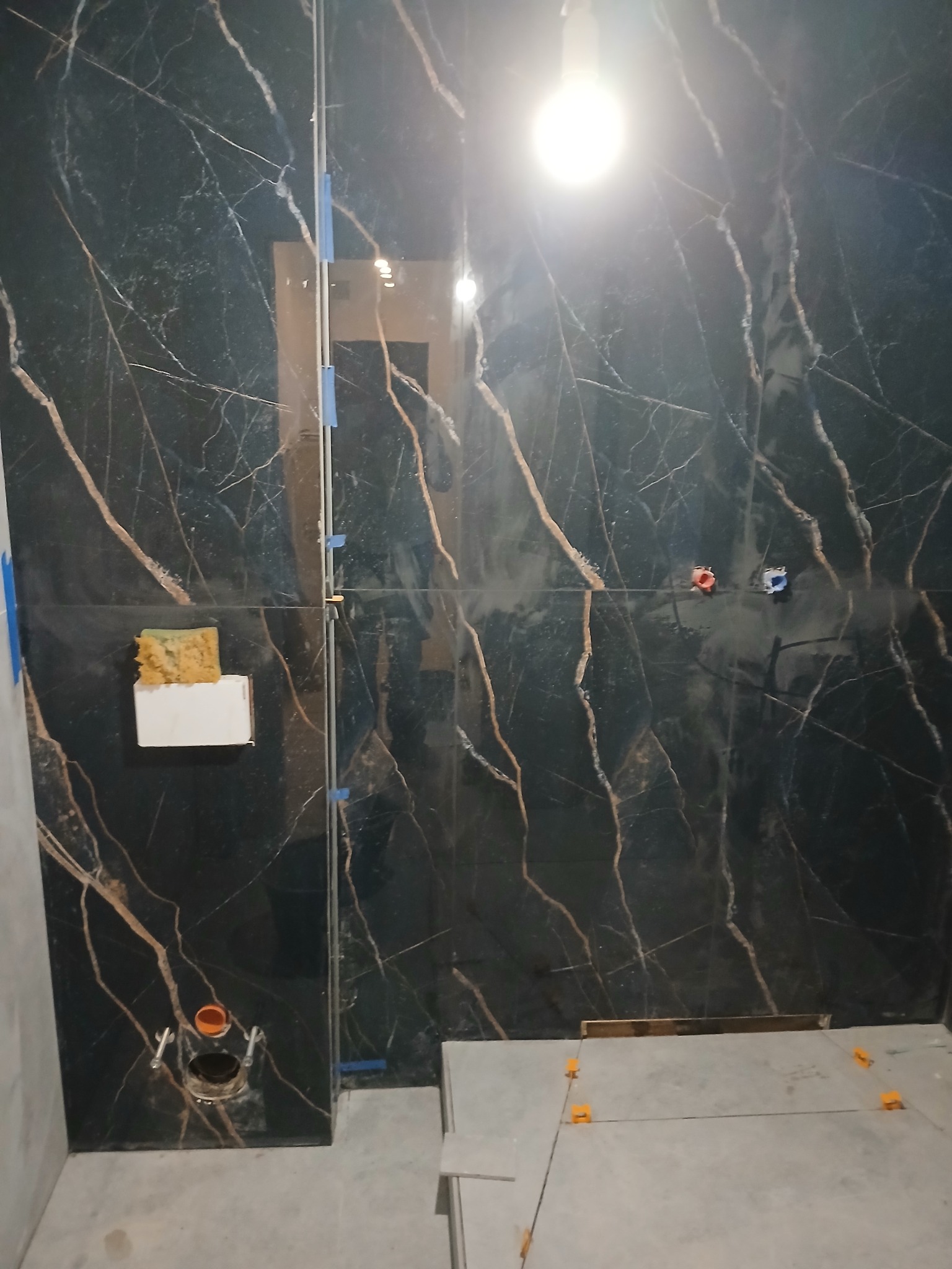 Częściowo wyłożona czarnymi płytkami z marmurowym wzorem ściana łazienki, widoczne otwory na armaturę, gąbka na płytce i poziomowanie płytek na podłodze.