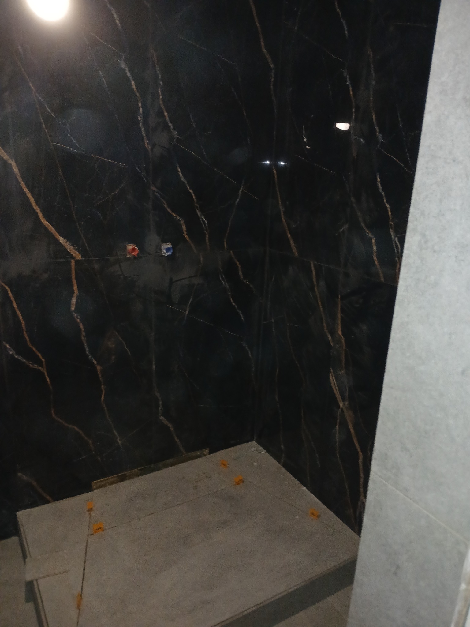 Montaż płytek: Czarna ściana z marmurowym wzorem i szare płytki podłogowe z pomarańczowymi klinami dystansowymi podczas układania w łazience.