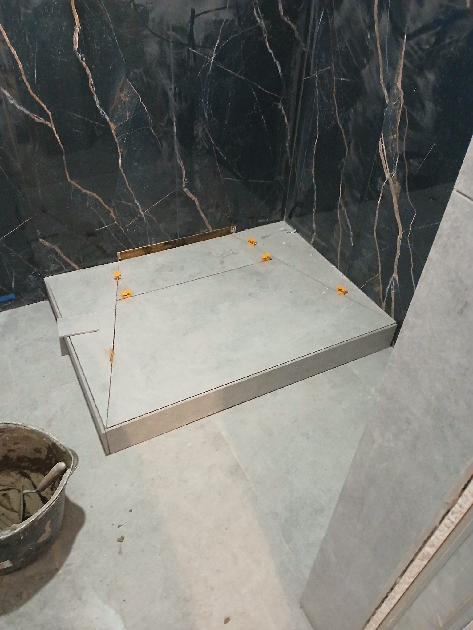 Montaż płytek podłogowych w kabinie prysznicowej z użyciem poziomowania krzyżykami, widoczna ściana z ciemnego marmuru z jasnymi żyłami i wiadro z zaprawą.