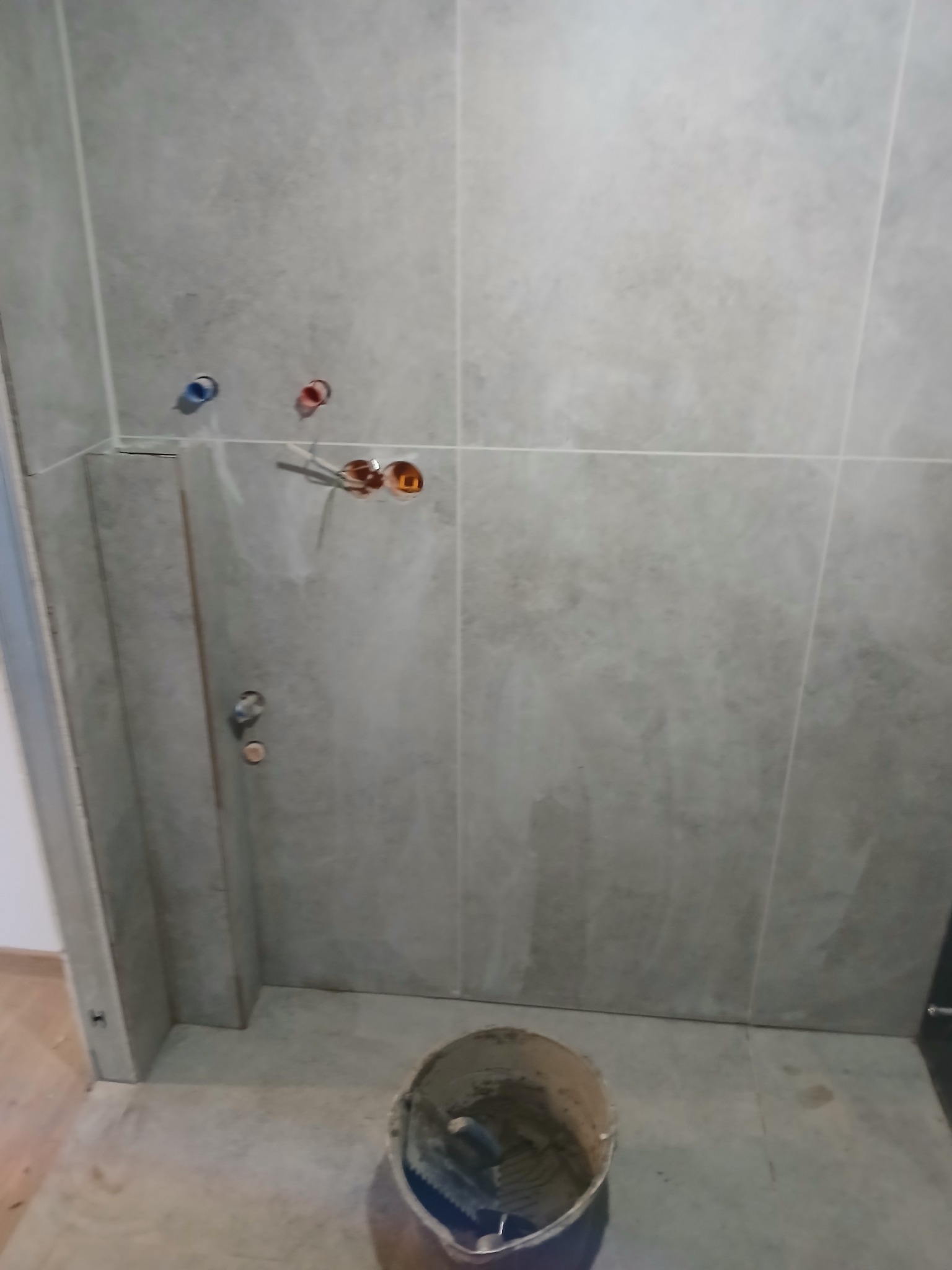 Szare płytki ścienne w łazience w trakcie montażu, widoczne rury z ciepłą i zimną wodą oraz wiadro z zaprawą klejową na podłodze.