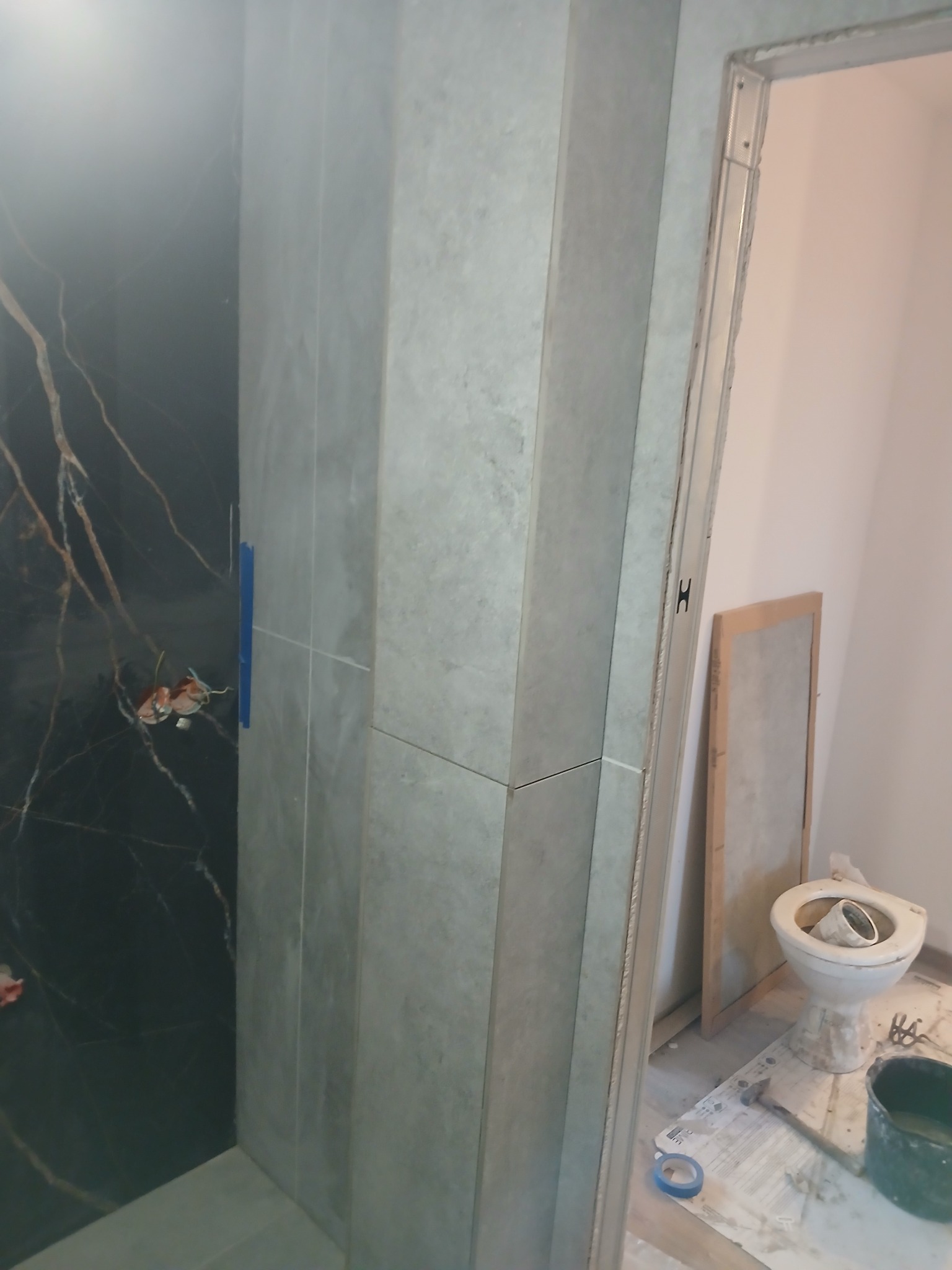 Wnętrze łazienki w trakcie remontu: częściowo wyłożone płytkami imitującymi czarny marmur z żyłami, szare płytki na ścianie, widoczna instalacja elektryczna zabezpieczona taśmą, nieukończona...
