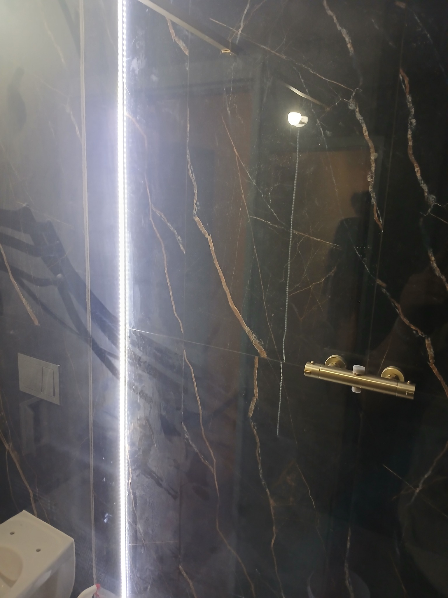 Część wykończonej łazienki z czarnymi płytkami imitującymi marmur, podświetlona pionową listwą LED, widoczna bateria prysznicowa w kolorze złotym oraz fragment toalety.