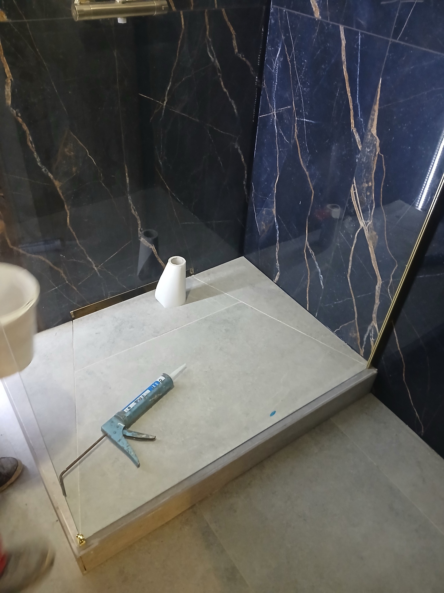 Szczegółowe ujęcie montażu kabiny prysznicowej z użyciem silikonu, widoczna aplikator silikonu, biała ceramika i czarne marmurowe płytki na ścianie.