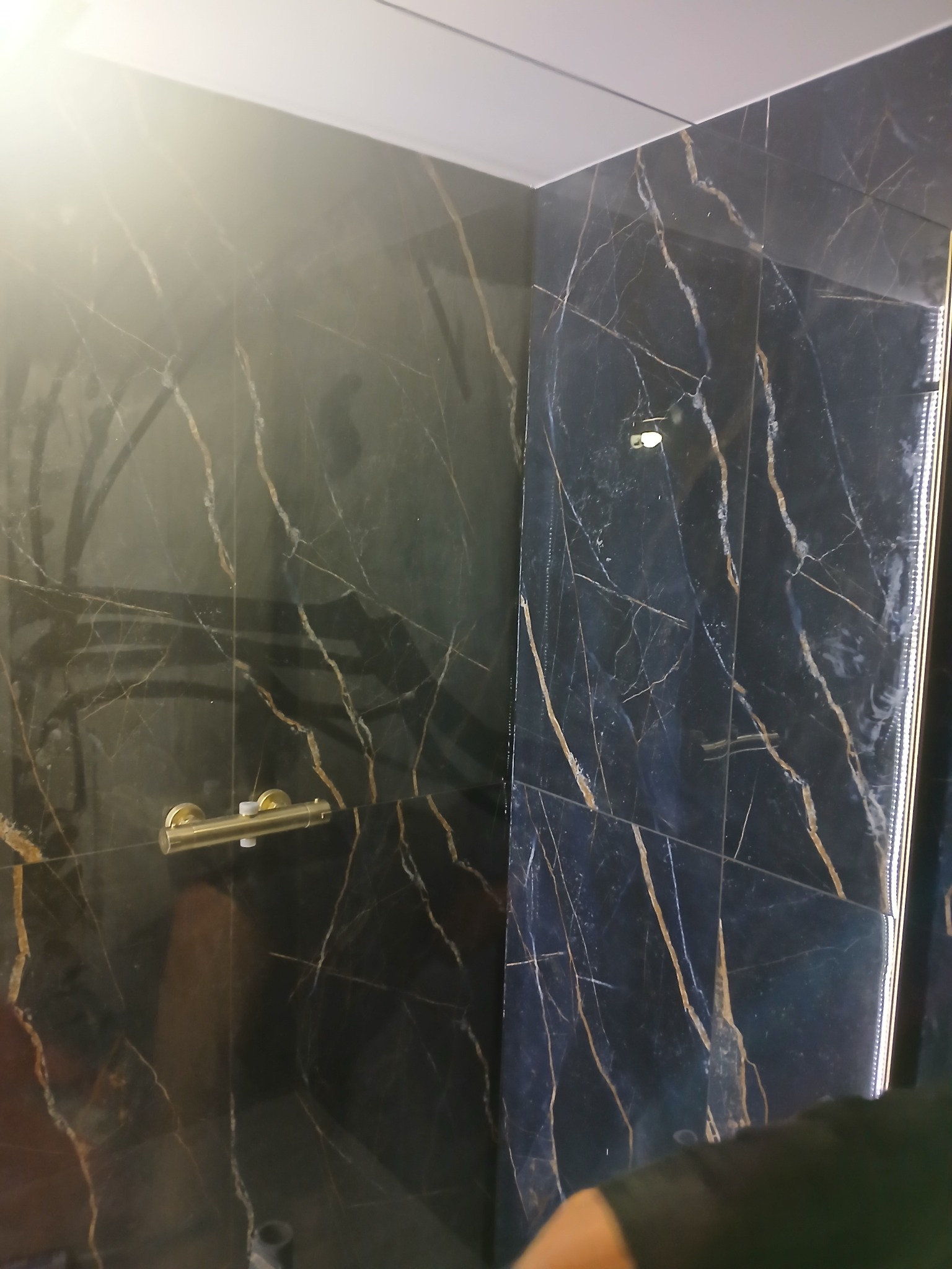 Ciemna, marmurowa ściana kabiny prysznicowej z złotymi żyłkami, widoczny złoty kran i oświetlenie LED w rogu.