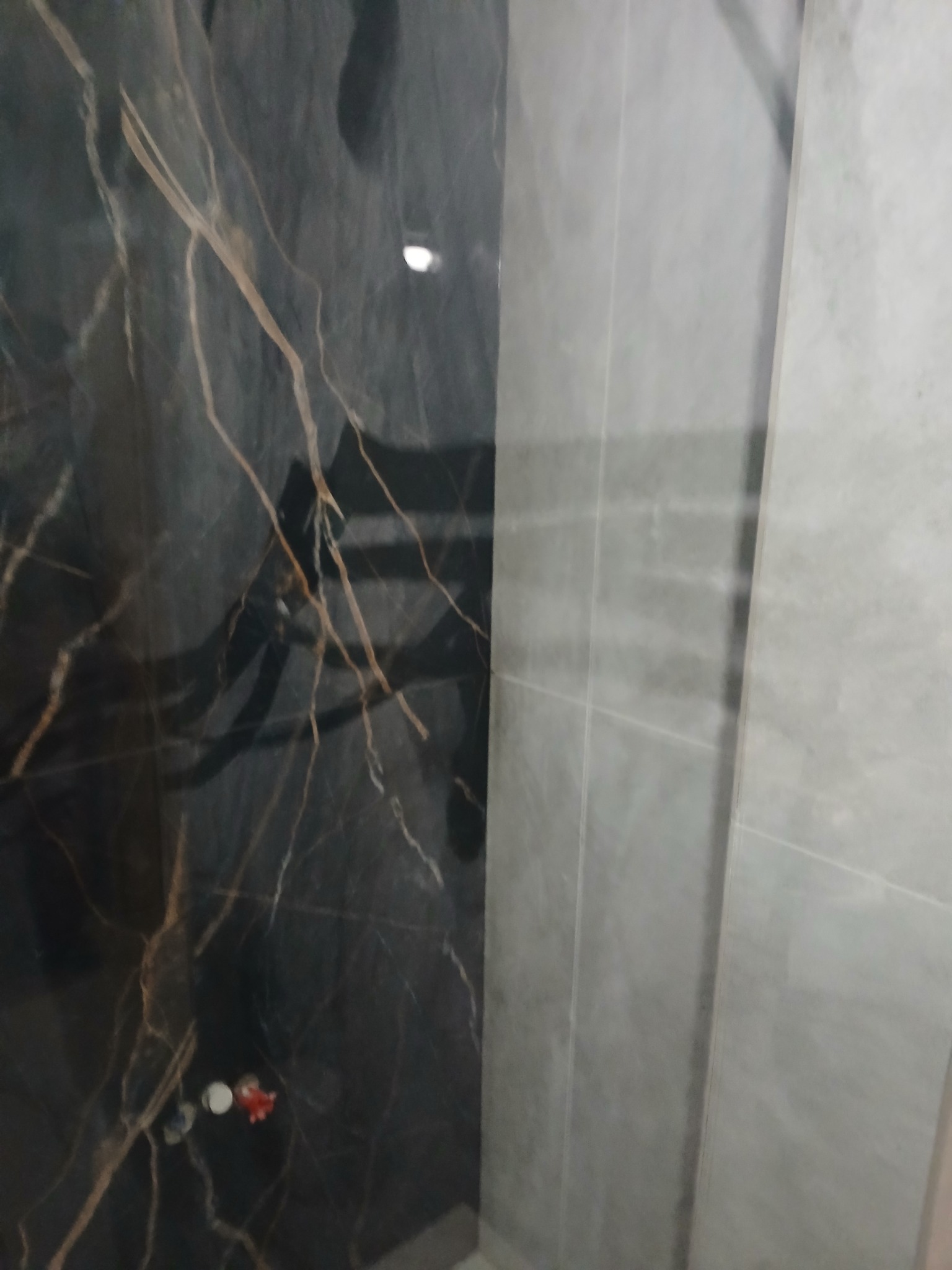 Częściowo wykończona łazienka z czarną płytką imitującą marmur z brązowymi żyłami po lewej stronie i szarą płytką po prawej, widoczne elementy instalacji wodnej.