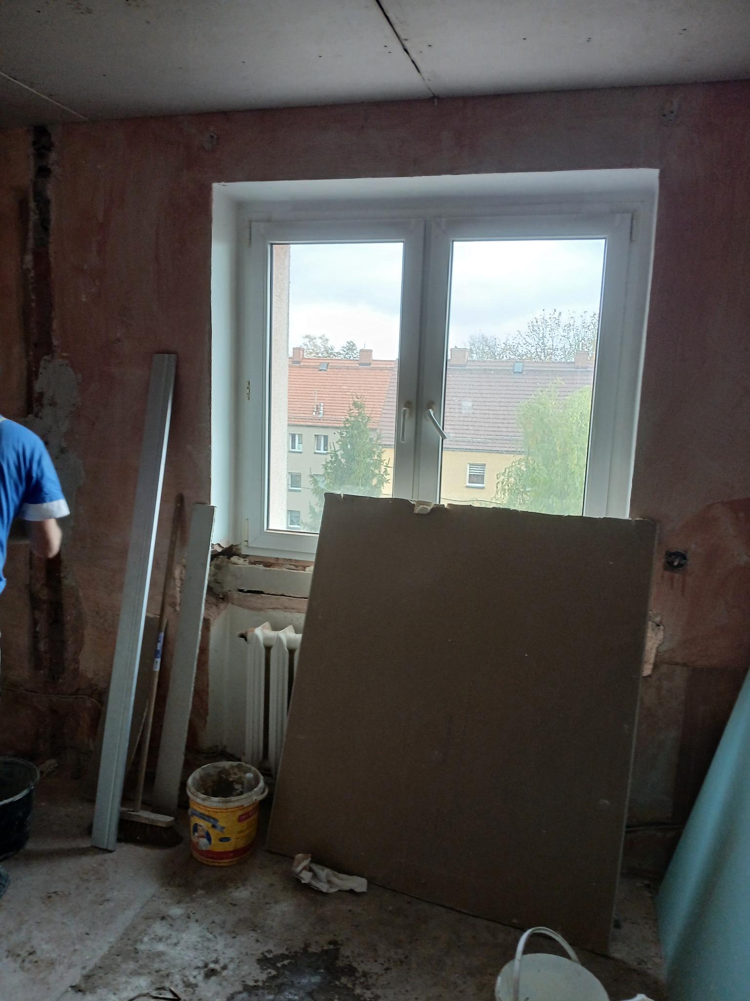 Pokój w trakcie prac remontowych: ściany z surowym tynkiem, okno z widokiem na budynki, płyta kartonowo-gipsowa oparta o ścianę, wiadro z farbą i listwy przypodłogowe.