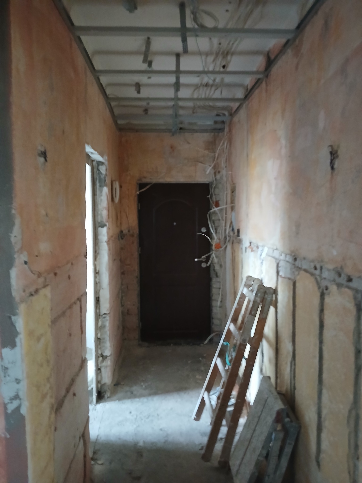 Korytarz w trakcie remontu: widoczne ściany przygotowane do wykończenia, metalowa konstrukcja pod sufitem, wystające przewody elektryczne, drabina oparta o ścianę.