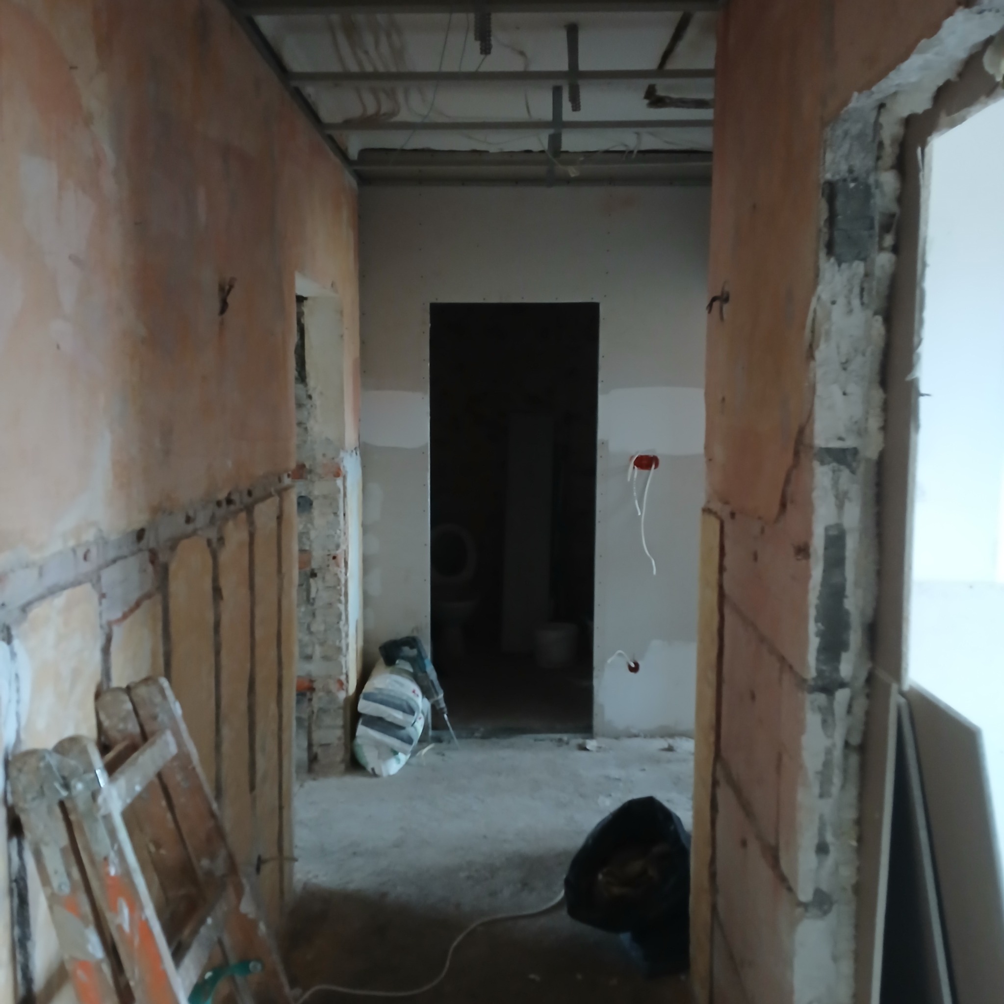 Korytarz w trakcie remontu: ściany częściowo otynkowane, widoczne instalacje elektryczne, drabina malarska i worki z materiałami budowlanymi. W tle widoczna toaleta.