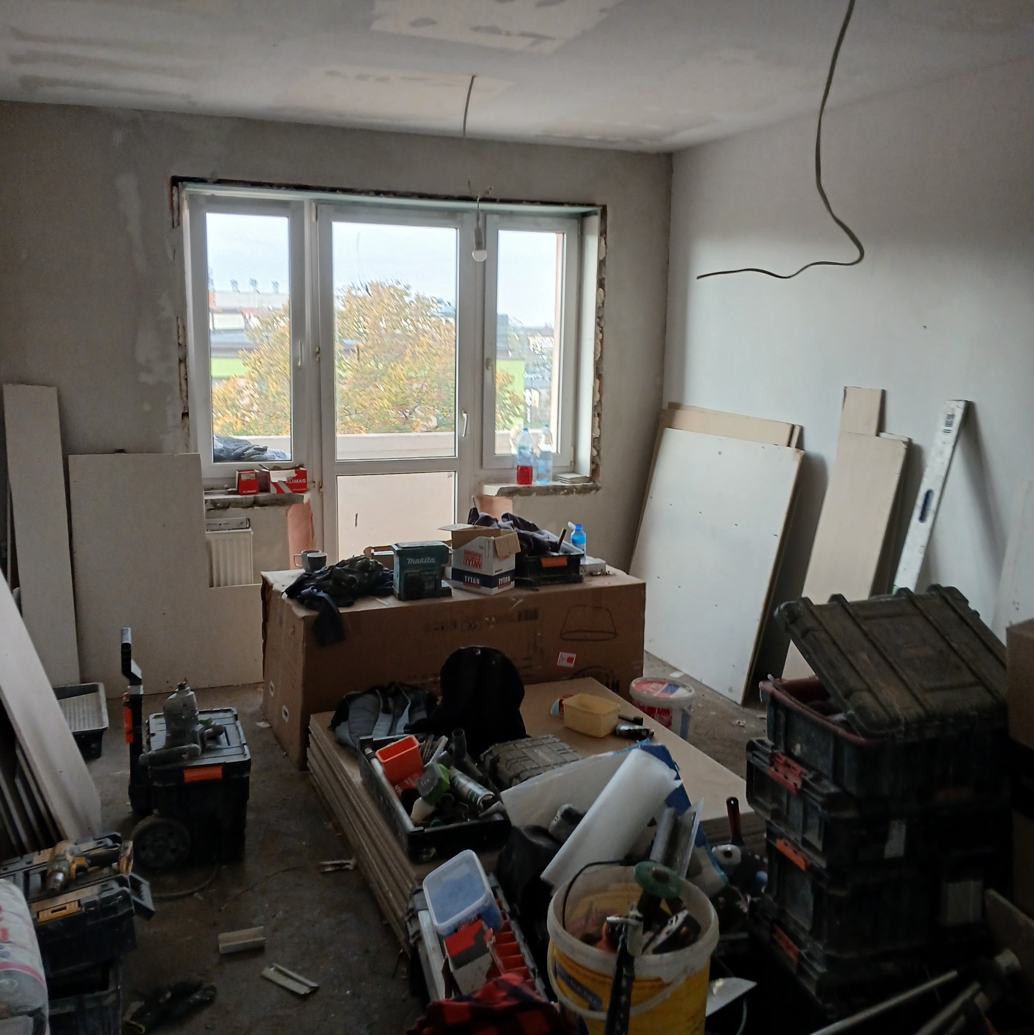 Pokój w trakcie remontu: widoczne narzędzia, płyty karton-gips, niezabezpieczone okno balkonowe, surowe ściany i wystające kable elektryczne.