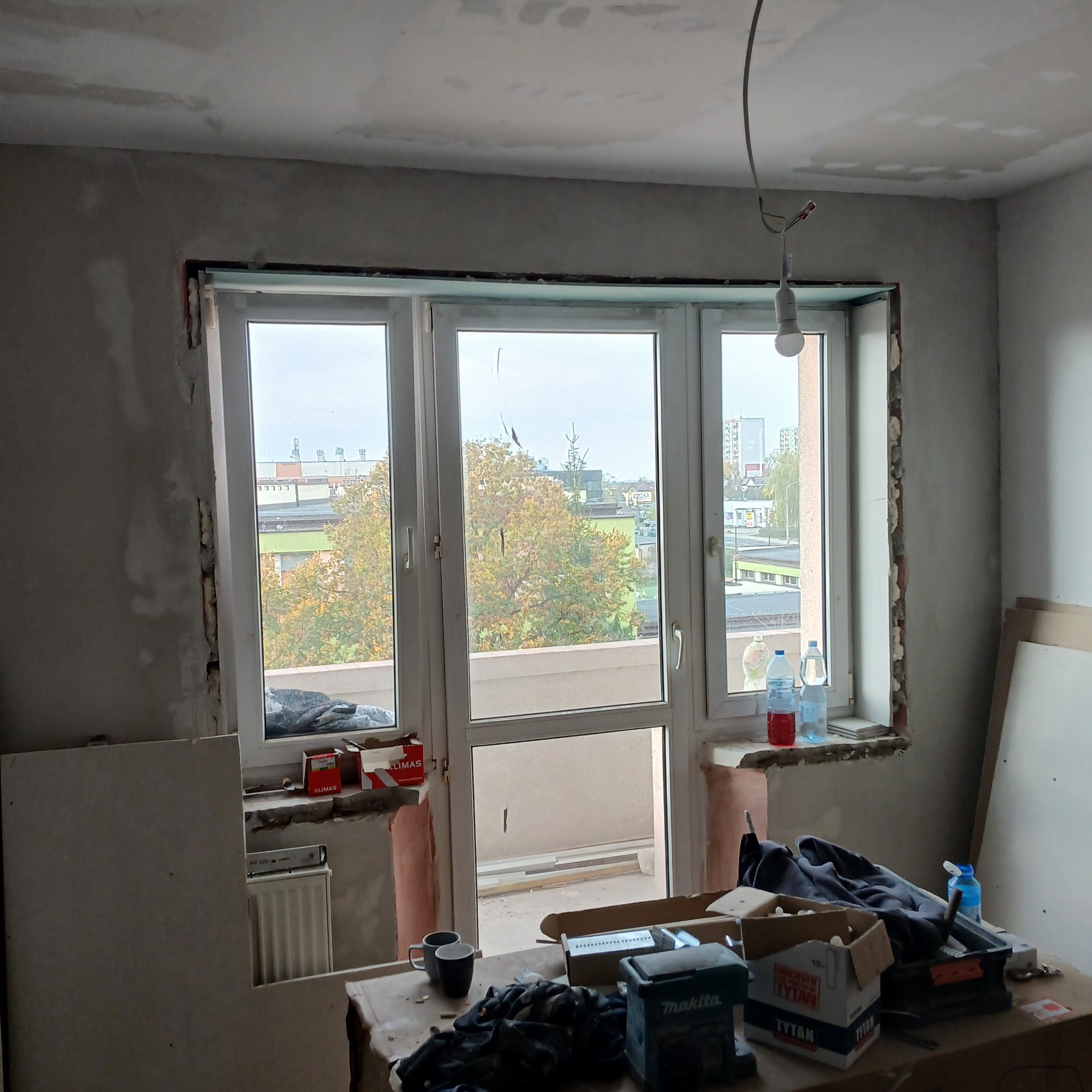 Pokój w trakcie prac wykończeniowych: surowe ściany wokół okna balkonowego, widoczne ślady po skuciu tynku, przygotowane płyty karton gips, narzędzia i materiały budowlane na prowizorycznym stole.