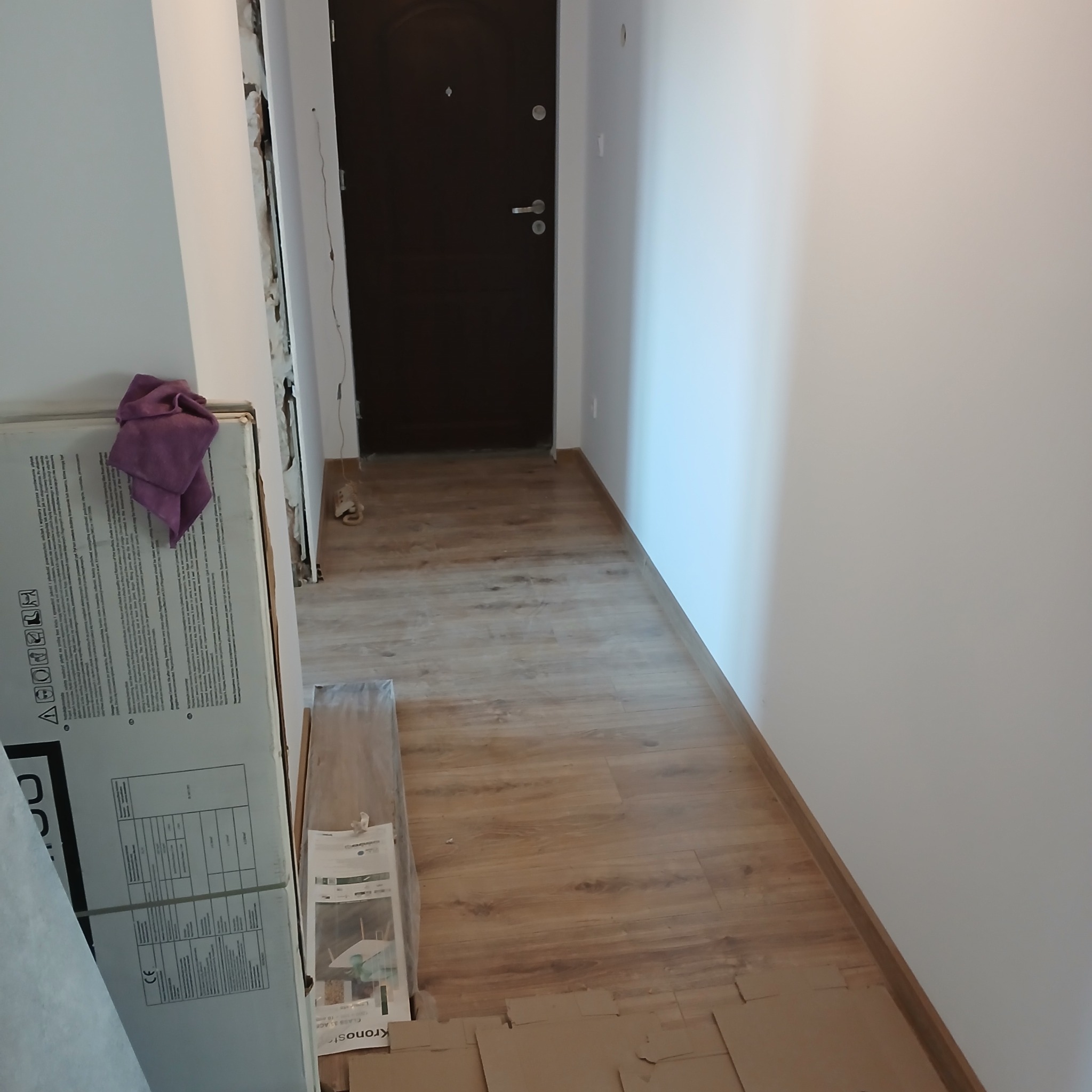 Korytarz w trakcie remontu: świeżo położona podłoga laminowana, zabezpieczona kartonem, białe ściany, ciemne drzwi wejściowe, karton z panelami podłogowymi i fioletowa ścierka w rogu.