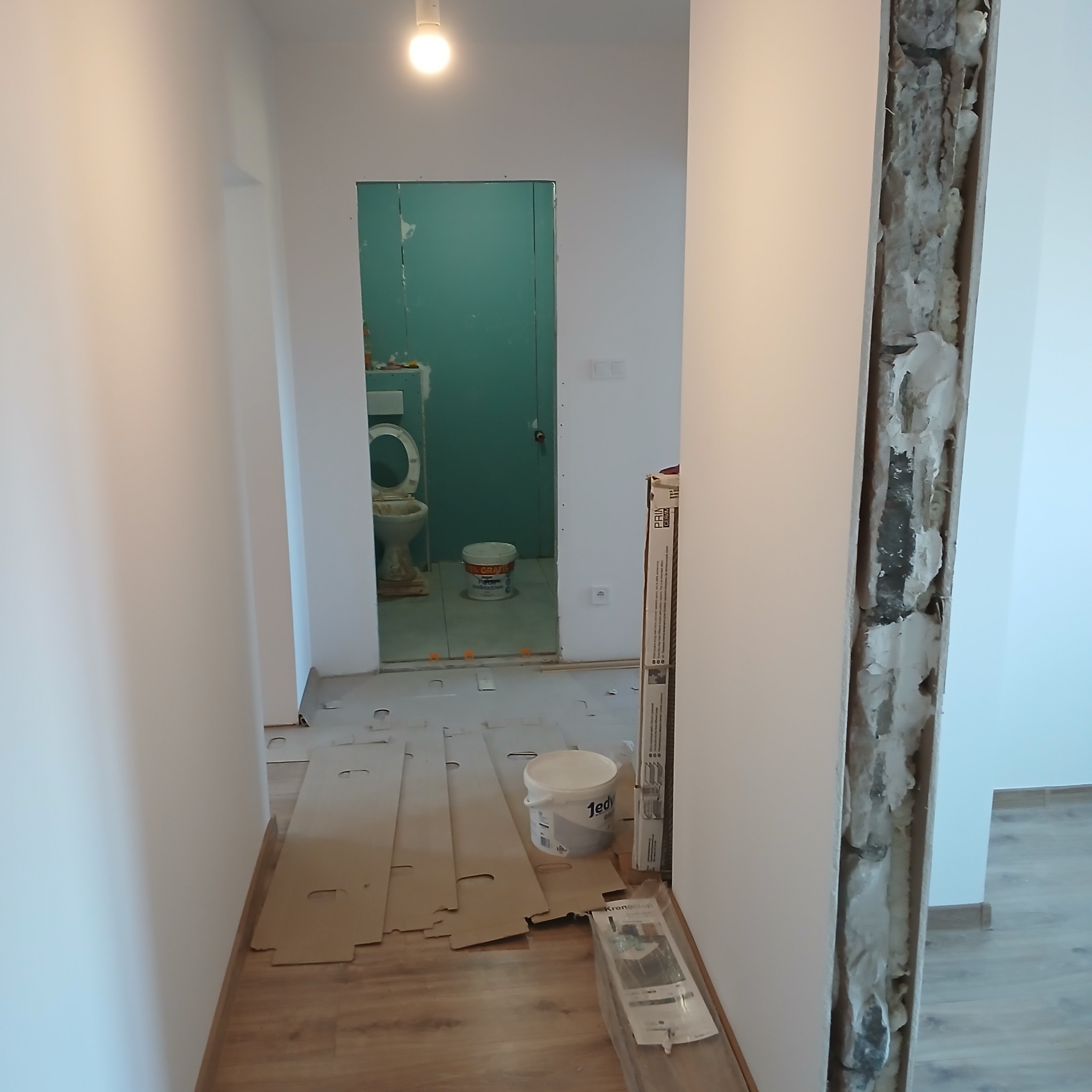 Korytarz w trakcie remontu z widoczną łazienką w tle, zabezpieczona podłoga, ściana z odsłoniętą izolacją, przygotowania do prac wykończeniowych.
