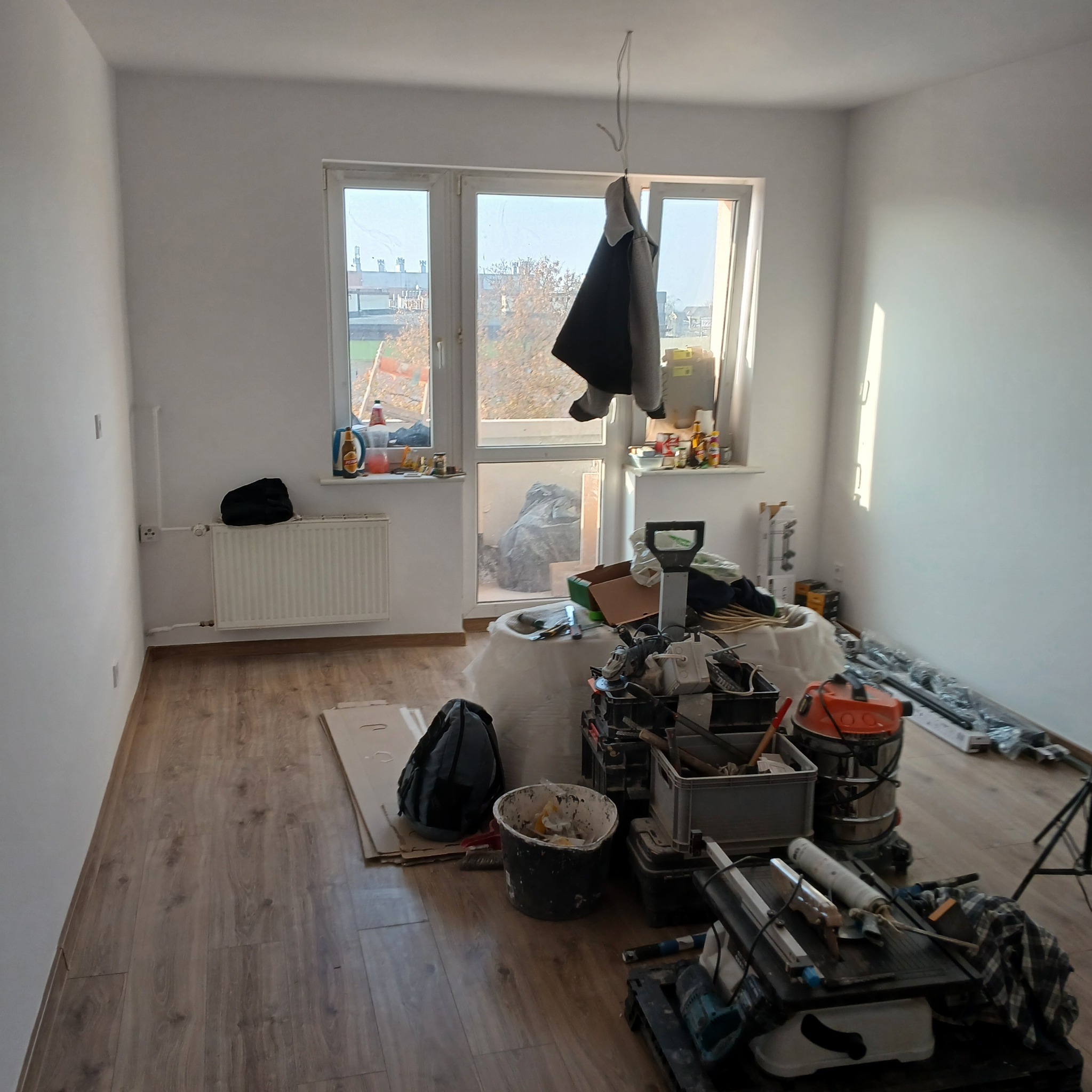 Pokój w trakcie remontu: świeżo pomalowane ściany, widoczne narzędzia, wiadro z farbą, odkurzacz przemysłowy, torba robocza, okno z widokiem na budynek i drzewa, ubranie wiszące na sznurku przy oknie.